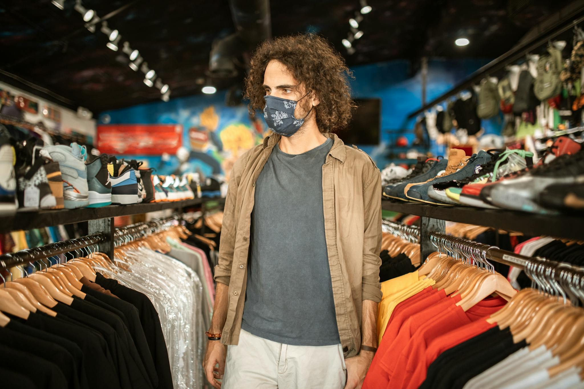 Man with curly hair wearing face mask shopping in a vibrant and trendy clothing store.